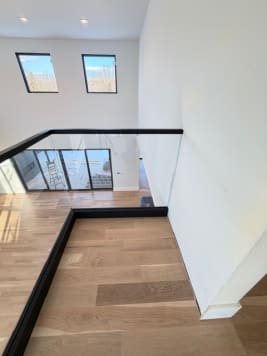 Frameless glass railing in a black base rail with black cap rail edges a white oak landing overlooking a great room.