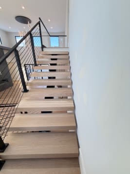 White oak floating treads rise along a white wall beside a black cable railing.