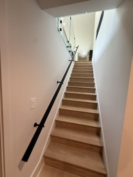 A black wall-mounted handrail runs up a long white oak stair with glass railing at the top.