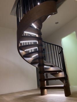 Dark oak wood-wrapped spiral stair with black metal railing and LED strips under each tread.