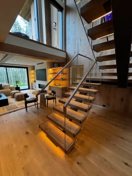 Oak floating treads glowing with under-tread LED light climb past a frameless glass guard with a wood handrail into an open living room.