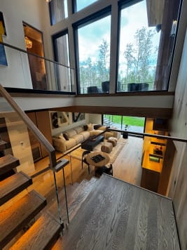 View from the upper landing past a frameless glass guard with wood handrail down into a bright double-height living room.