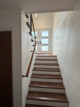 A wood handrail on black brackets runs along a white-paneled stairwell leading to a glass front door.