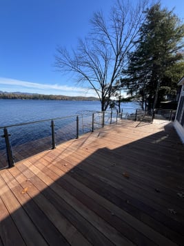 Black posts hold glass panels along an ipe deck overlooking a lake with autumn trees.