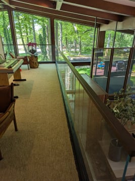 A long frameless glass guard with wood cap lines a loft walkway beside floor-to-ceiling windows facing woods.