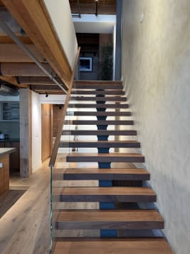 Dark walnut floating treads on a blue center stringer rise beside a textured plaster wall with frameless glass railing in a timber-beamed loft.