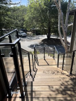 Looking down a wide wood exterior stair lined on both sides with black post glass railing toward a wooded driveway.