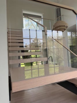 White-oak floating treads rise behind frameless glass railing with an oak handrail in front of tall grid windows.