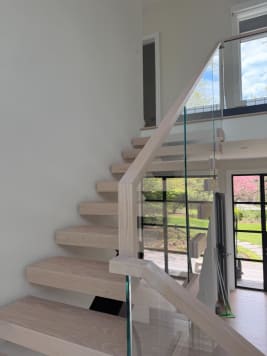 White-oak floating treads climb beside frameless glass with an oak handrail toward a bright window wall.