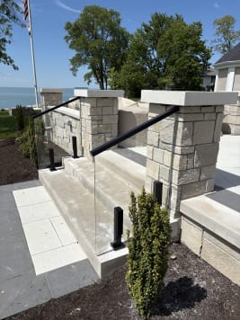 Frameless glass in black talon clamps with a black handrail flanks limestone steps between stone pillars near a lake.