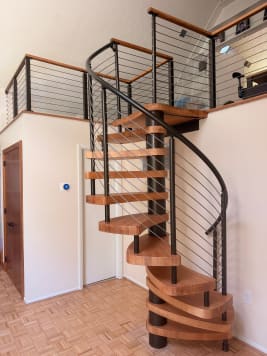 Light wood spiral stair on a black center column with curved cable railing up to a cable-railed loft.