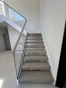 Gray-washed oak floating treads rise beside frameless glass in a white base rail with a matching wood handrail.