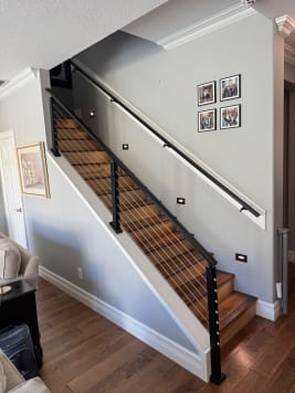 A black wall-mounted metal handrail opposite black cable railing on a stained wood stair.