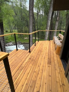 Cedar deck corner with black-post cable railing and cedar cap overlooking rapids and cottonwood trees.