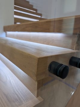 Close detail of black standoff pins holding glass to thick oak treads with under-tread LED lighting.