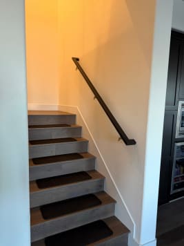 Black wall-mounted handrail along a closed stair with gray treads and dark tread mats.