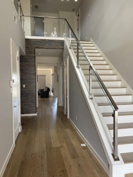 Stainless posts with horizontal rod infill and a matching handrail run up a white straight stair beside a long wood-floored hallway.