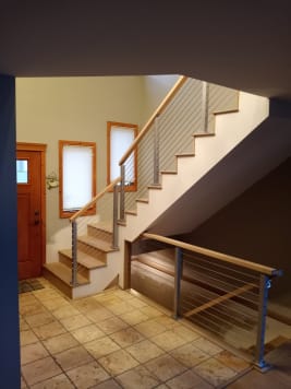 Cable railing with a wood handrail climbs a white stair and guards the opening beneath it beside a wood entry door.