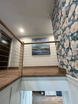 Oak newels and cap rail with cable infill around a stair opening between white wainscoting and floral wallpaper.
