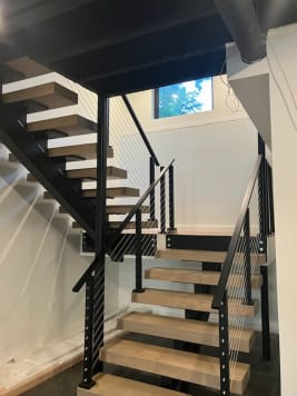 Thick light oak treads on a black mono stringer with black cable railing rising toward a window.
