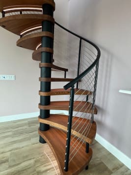 Spiral staircase with wood treads on a black center column and a curved black handrail with stainless cable infill, in a bright room with tile floors.