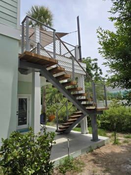 Side view of an exterior FLIGHT mono stair with hardwood treads and silver cable railing climbing to a raised deck among tropical plants.
