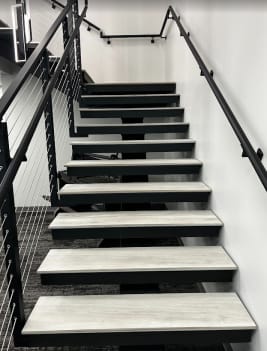 Gray treads on a black mono stringer between black cable railing and a wall-mounted black handrail in an office stairwell.