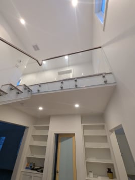 Frameless glass panels on stainless standoff pins with a wood handrail line a white stair and upper balcony.
