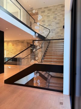 A black-framed floating stair with glass railing rises through a stone-walled atrium past a glass-floored landing.