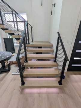 White oak floating treads with LED underglow and black cable railing on both sides in a modern stairwell.