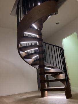 Dark oak wood-wrapped spiral stair with black metal railing and LED strips under each tread.
