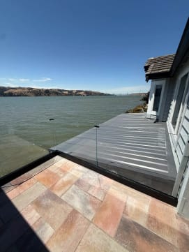 Frameless glass in a black base channel edges a tiled waterfront patio overlooking a wide bay and hills.