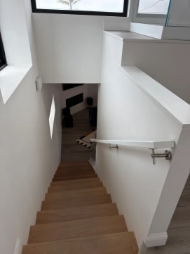 A slim white handrail on stainless brackets runs down a narrow oak stair between white walls.
