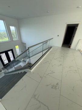 Frameless glass with a gray top cap rings a stair opening on a white marble-tile floor below tall windows.