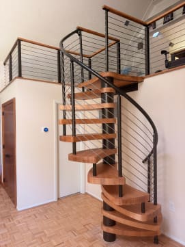 Light wood spiral stair on a black center column with curved cable railing up to a cable-railed loft.