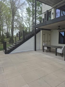 Dark composite stair with cable railing climbs from a covered patio to an upper deck among trees.
