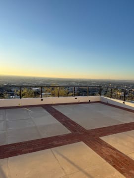 Cable railing tops a low wall around a brick-banded rooftop terrace with a sunset city view.