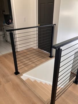 Black rod railing wraps the top of a stair opening on a light wood floor in a hallway.