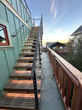 Slatted exterior treads with soft LED glow climb a sage-green wall beside black cable railing at sunset.