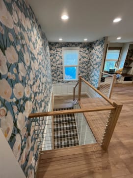 Oak newel posts and cap rail with stainless cable infill around a carpeted stair opening with floral wallpaper.