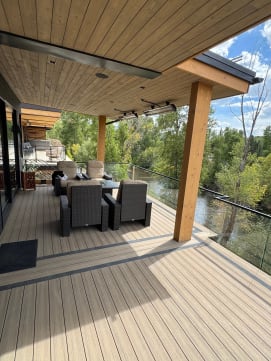 Frameless glass railing on black standoff pins surrounds a covered composite deck with lounge seating by a river.