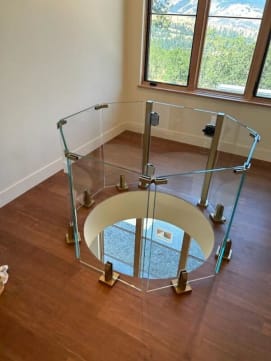 Faceted ring of frameless glass panels on posts guards a circular floor opening in a wood-floored room with mountain-view windows.