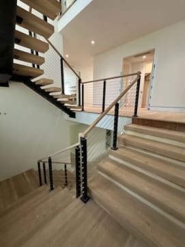 White oak treads on a dark stringer switch back at a landing, lined with black-post cable railing and a white oak handrail.