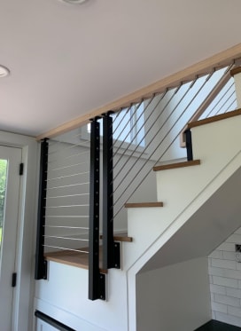 Black posts with angled cable infill fill the triangular opening beneath a white stair with a wood cap.