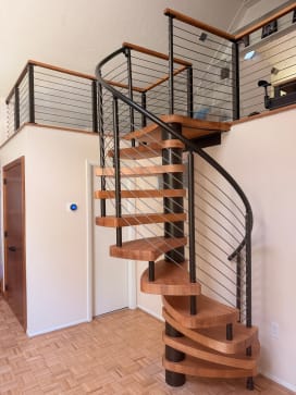 Light wood spiral stair on a black center column with curved cable railing up to a cable-railed loft.