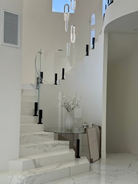 Black talon clamps hold curved glass along a white marble stair with a glass-top table at its base.