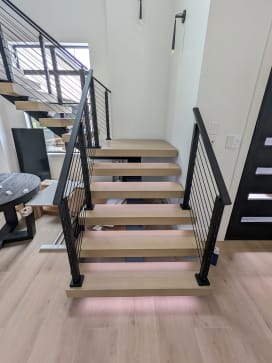 White oak floating treads with LED underglow and black cable railing on both sides in a modern stairwell.