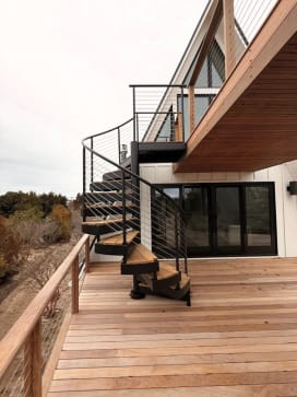 Black steel spiral stair with Ipe-wrapped treads and cable railing climbs from a wood deck to an upper balcony.