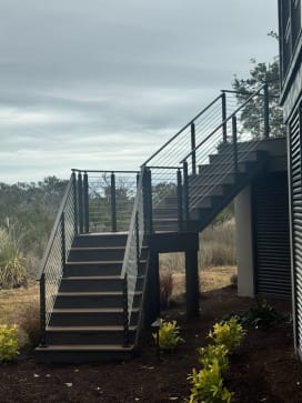 Black posts with horizontal rod infill follow a composite exterior stair and landing up to a raised coastal deck.
