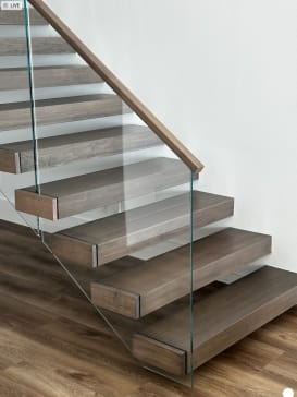 Thick gray-brown floating treads project from a white wall with frameless glass and a wood handrail.